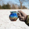 a hand holding a small round ceramic mug in brown yellow and blue glaze, with snow in the background