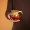 a hand holding a small round ceramic mug in green yellow and rustic red glaze
