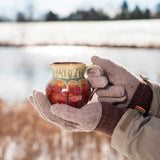 Two hands holding a handmade pottery coffee mug in orange red and green outside in the snow, great as a winter gift