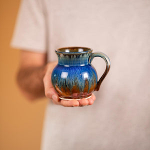 handmade small round pottery mug in amber blue glaze held by a person, holds ~11 oz of liquid