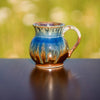 Handmade small pottery mug with a round shape and a handle, with brown and blue drips, sitting on a table