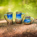 3 handmade pottery coffee mugs in brown yellow and blue glazes sitting out on the ground with a creek behind
